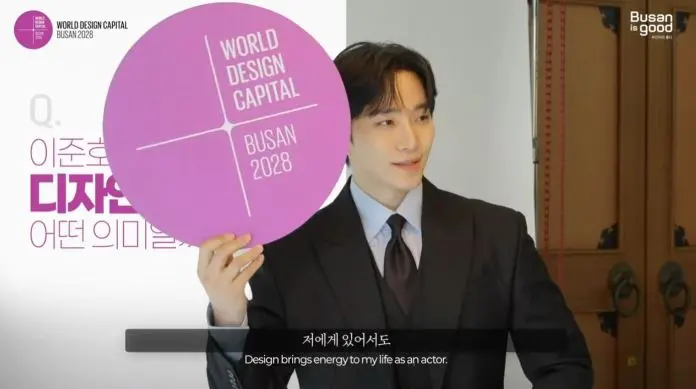 Lee Junho  in a formal suit holding a circular sign that reads 'WORLD DESIGN CAPITAL BUSAN 2028', with text in the background discussing the significance of design.