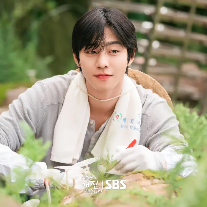 Ahn hyo seop with dark hair sitting in a garden, wearing a gray shirt and white gloves, tending to plants with a small tool.