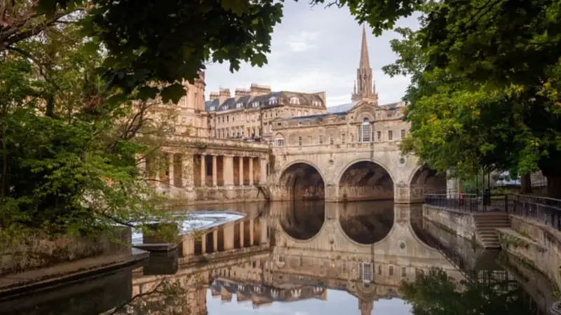 A view of Bath in the UK at sunrise featuring a river, bridge, and steeple framed by trees