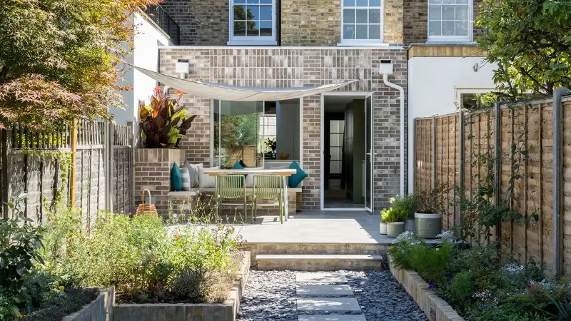 a narrow terrace garden with L-shaped paved garden path and a deck with table and chairs in the sun, and planter boxes running along the fenceline