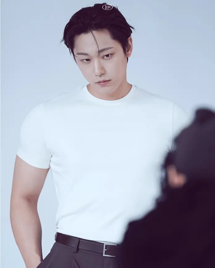 Lee Do hyun with short dark hair wearing a white t-shirt and black trousers, looking down thoughtfully against a plain background.