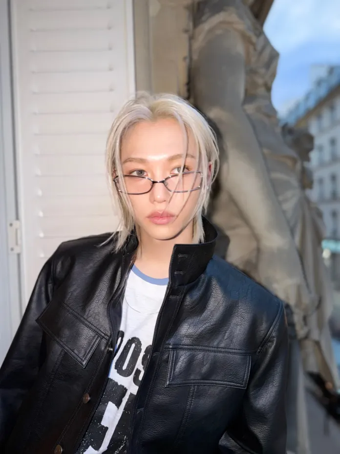 Felix with long platinum blonde hair wearing glasses and a black leather jacket, posed in front of a window with a statue in the background.