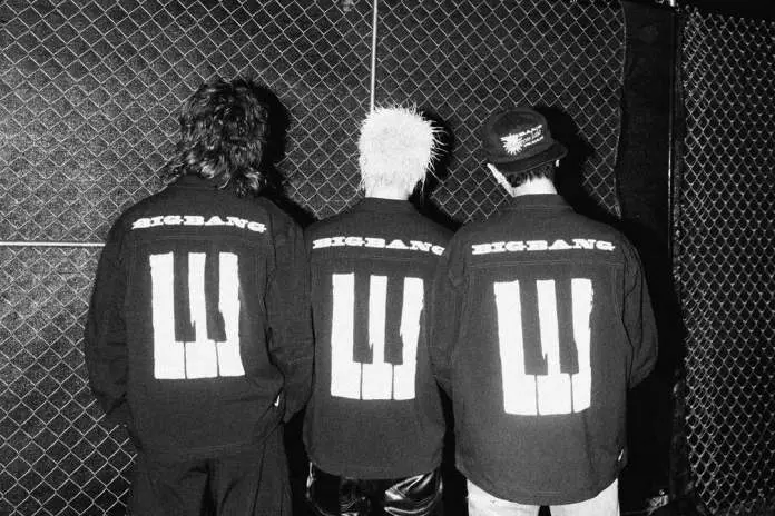 BigBang members standing with their backs facing the camera, wearing black jackets with 'BIGBANG' printed on the back and a piano key design.