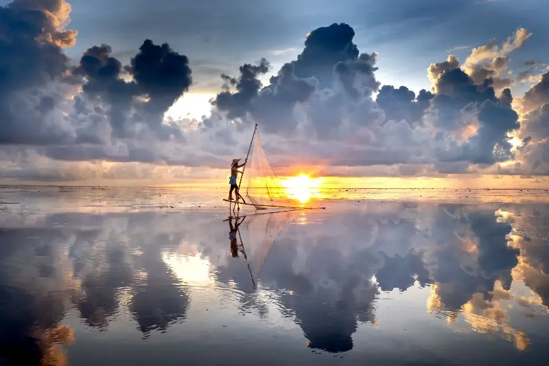 Fisherman casts net at sunrise, creating a serene reflection on calm waters.