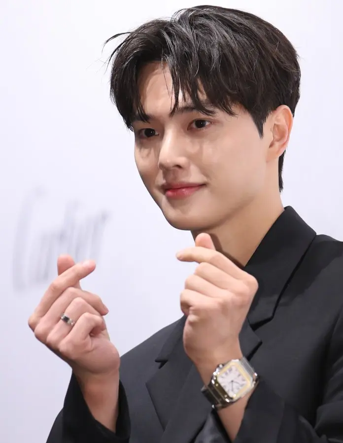 Song Kang with styled dark hair smiles and makes finger heart gestures, wearing a black suit and a silver wristwatch.