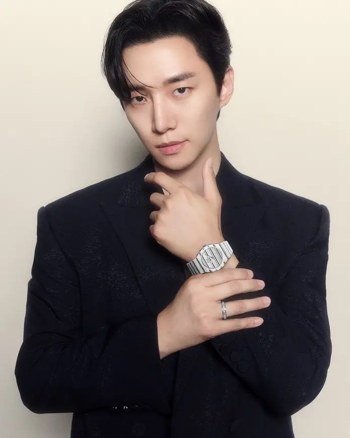 A person wearing a black blazer poses confidently, showcasing a watch on their wrist while touching their chin with one hand.