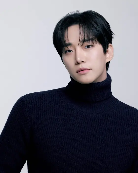 A young man with dark hair wearing a navy turtleneck sweater, looking at the camera against a plain background.