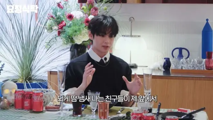 BTS V sitting at a table adorned with flowers and drinks, gesturing while speaking.