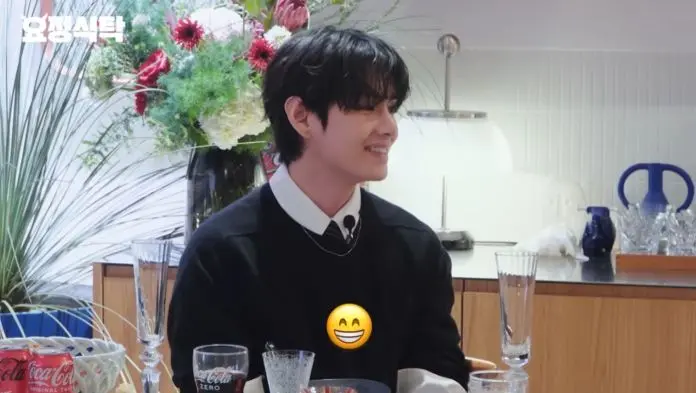 BTS V with dark, wavy hair is smiling while seated at a table adorned with flowers and various drinkware.