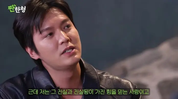Lee Min-ho with short black hair wearing a leather jacket, speaking thoughtfully in a seated position, with a blurred background.