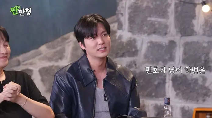 Lee Min-ho in a black leather jacket is sitting and speaking with an emotional expression, while another man in a black shirt is smiling beside him. The background features a stone wall and a bottle on the table.