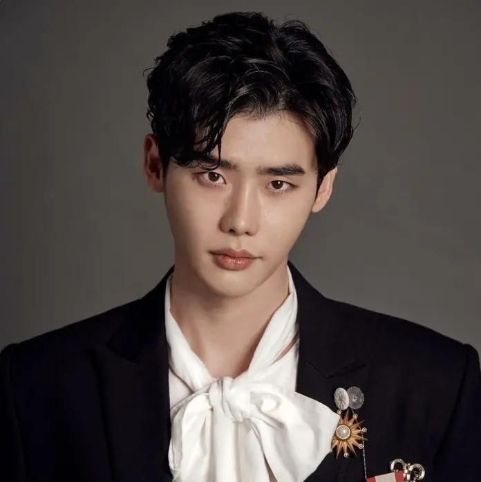 Lee Jong Suk with dark, styled hair, wearing a black suit jacket with decorative pins and a white scarf tie, looking directly at the camera.