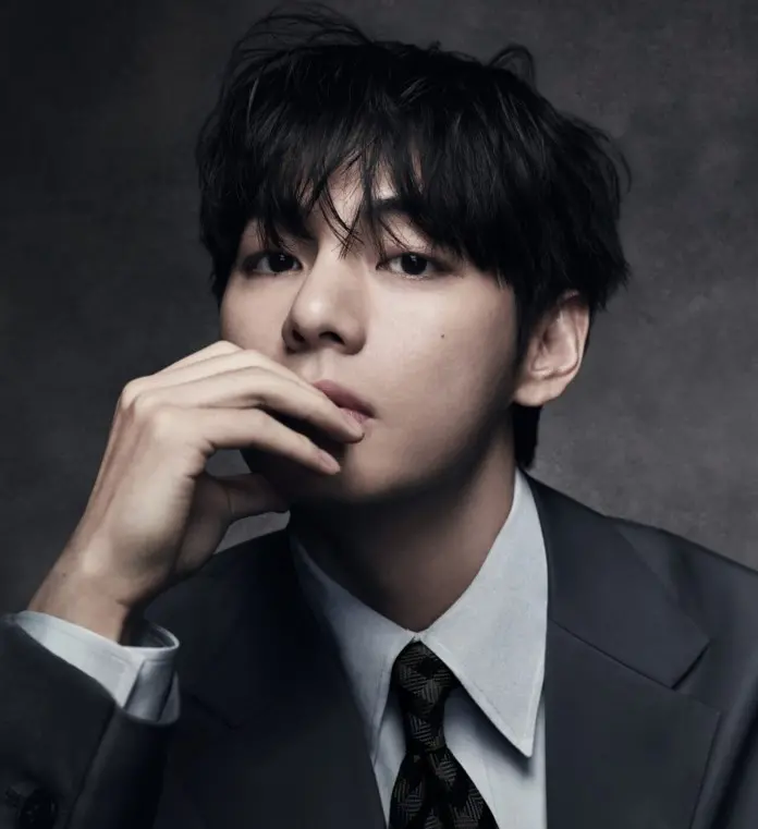 A close-up portrait of a young man with dark, tousled hair, wearing a formal suit and tie, resting his chin on his hand and gazing thoughtfully at the camera against a dark background.