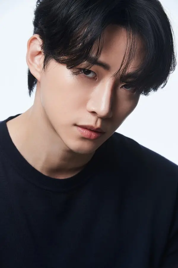 Close-up portrait of Lee Junho with dark hair, wearing a black shirt, against a light background, looking directly at the camera.