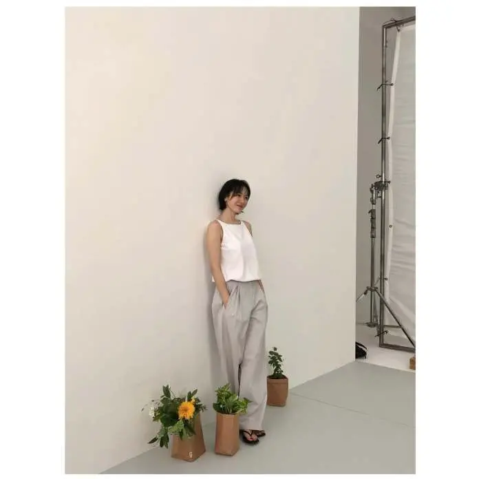 Son Ye jin in a white tank top and light gray pants stands against a plain wall, smiling casually. Two brown paper bags with plants and flowers are placed at her feet.