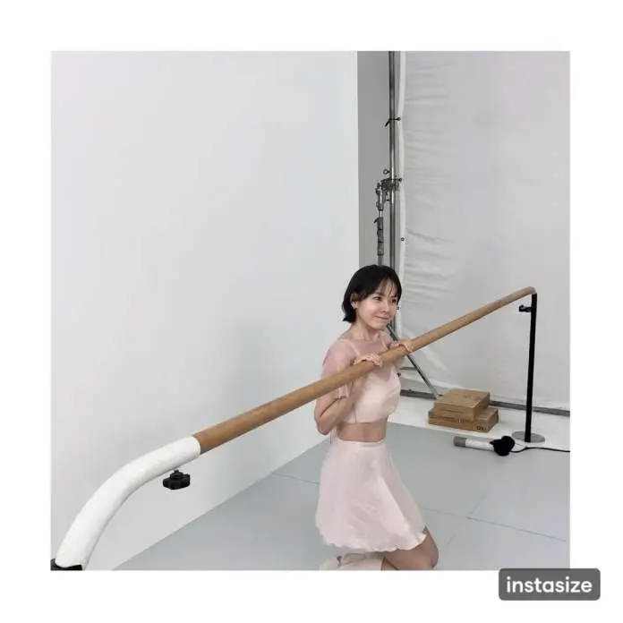 Son Ye jin posing in front of a simple studio backdrop, holding onto a wooden bar while dressed in a light pink outfit.