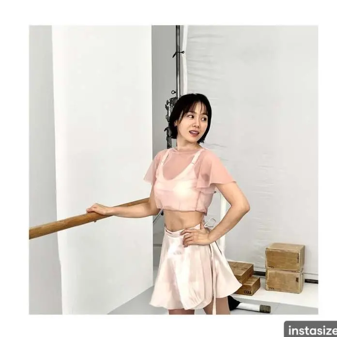 Son Ye jin posing in a studio, wearing a stylish pink outfit consisting of a cropped top with sheer sleeves and a matching skirt. She is standing near a wooden railing with studio equipment and boxes in the background.