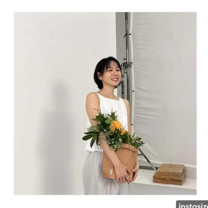 Son Ye jin smiling wearing a white tank top and light gray pants holds a bouquet of flowers in a tan bag, standing in a bright studio setting.