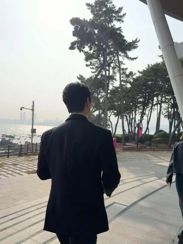 Lee Junho in a black coat walking away from the camera along a waterfront promenade surrounded by trees, with a hazy skyline in the background.