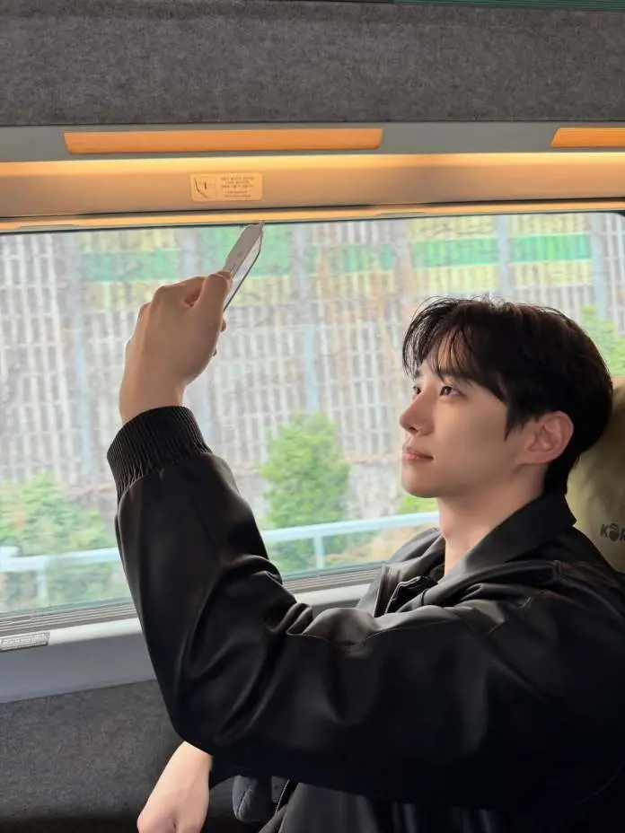 Lee Junho sitting in a train, holding a smartphone and taking a selfie, with a focused expression, while greenery is visible outside the window.