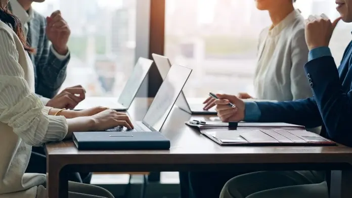 A group of professionals engaged in a meeting at a table, using laptops and taking notes, with a city skyline in the background.