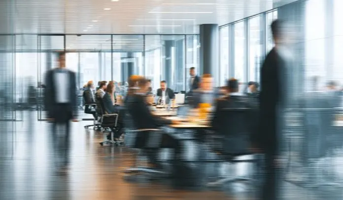 A blurred image of a modern office environment with people engaged in discussions around a conference table.