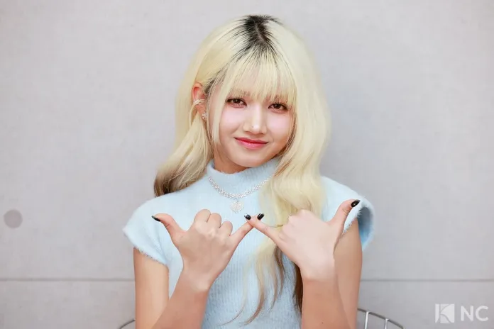 Hara with long, blonde hair wearing a light blue sleeveless top, making a hand gesture with both hands, smiling at the camera.