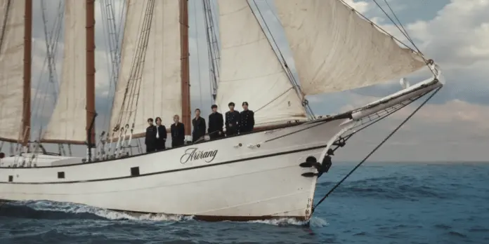 A sailing ship named 'arirang' with multiple sails, carrying BTS dressed in black uniforms, navigating through the ocean under a cloudy sky.