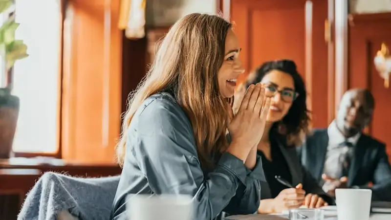 Smiling businesswoman with hands clasped communicating with colleagues in meeting room at office