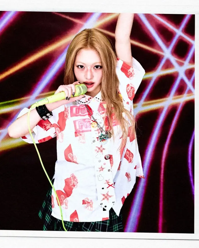 A young person with long blonde hair holds a green microphone while striking a pose. They wear a colorful shirt with graphic prints and a plaid skirt, set against a vibrant, abstract background with light beams.