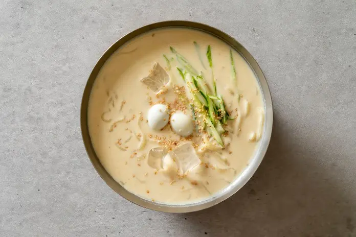 A bowl of creamy noodle soup topped with sliced cucumber, quail eggs, sesame seeds, and ice cubes on a light gray surface.