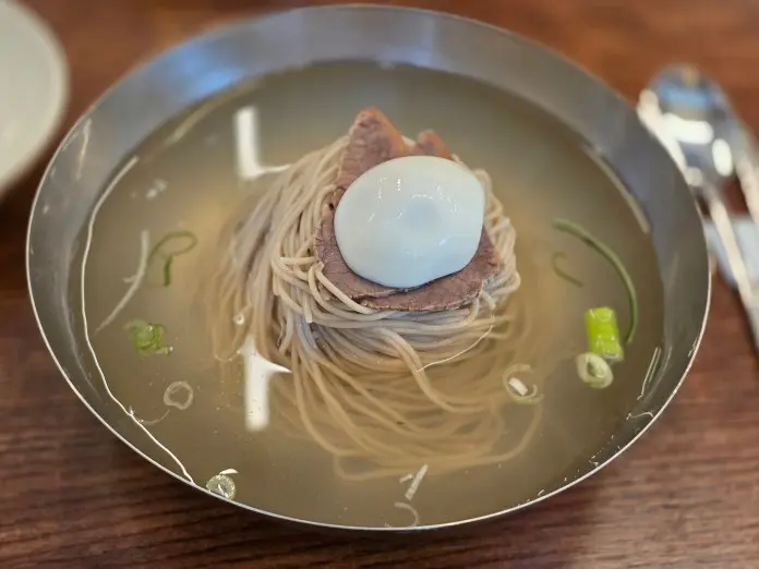 Bowl of cold noodles topped with a poached egg and slices of beef in clear broth, garnished with green onions.