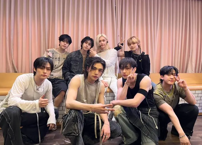 Stray Kids posing together, showcasing a stylish and casual fashion aesthetic. They are smiling and using hand gestures, with a background of curtains and a wooden surface.