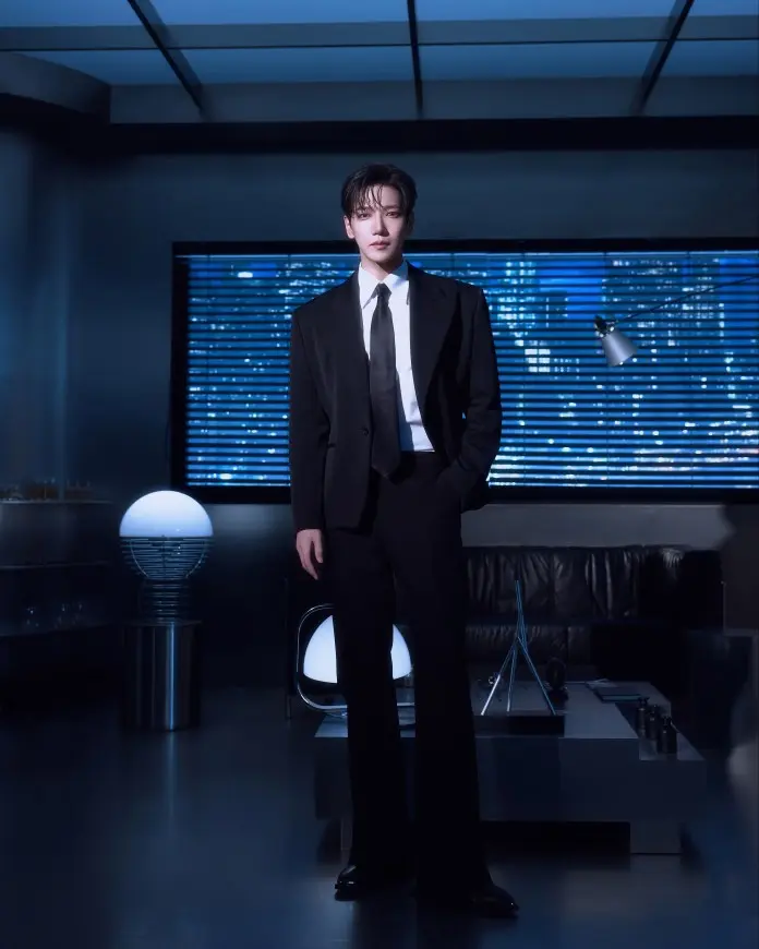 JUN. K in a black suit and tie stands confidently in a modern, stylish indoor space with blue lighting, featuring a leather sofa and contemporary decor.