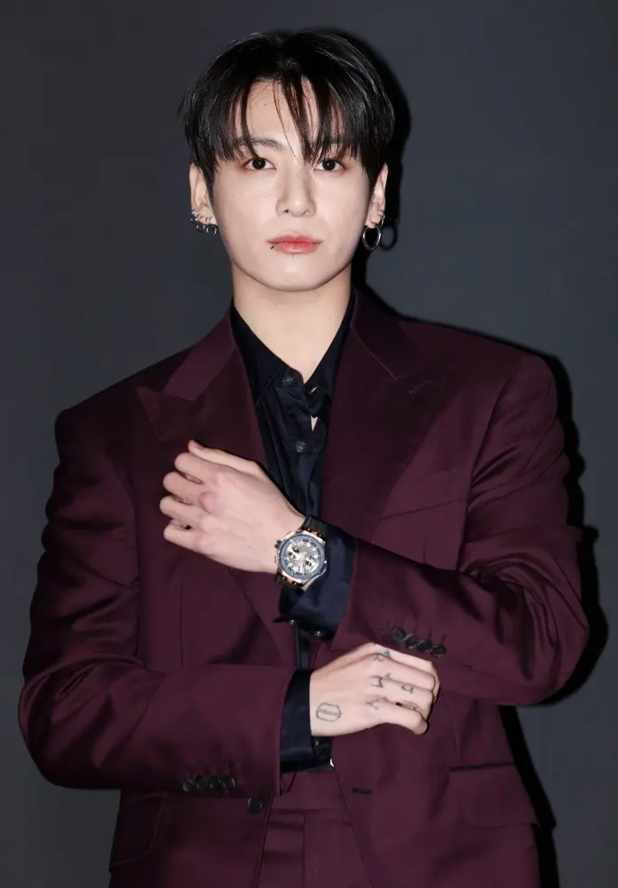 Jungkook in a maroon suit stands confidently with one hand on his arm, wearing a black shirt and a watch, against a dark background.
