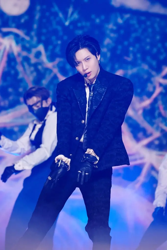 Taemin in a stylish black outfit and gloves stands confidently on stage, with a dramatic background and additional dancers in the scene.