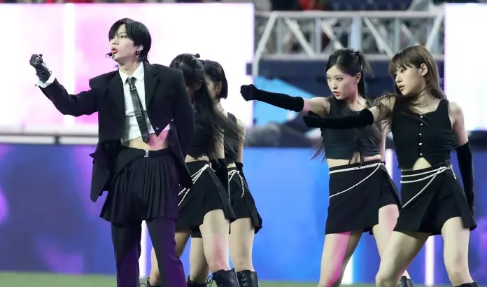Taemin in a stylish outfit sings passionately while surrounded by four female dancers in black attire, all engaged in a dynamic choreographed performance.