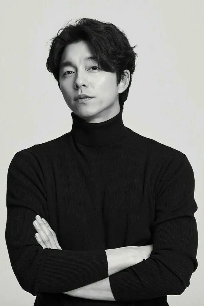 Gong Yoo with medium-length dark hair is posing confidently, wearing a black turtleneck sweater. He has his arms crossed and is looking directly at the camera against a plain white background.
