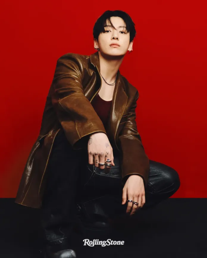 Jung Kook wearing a brown leather coat and black jeans, crouching against a red background, with a contemplative expression and visible tattoos on their hands.