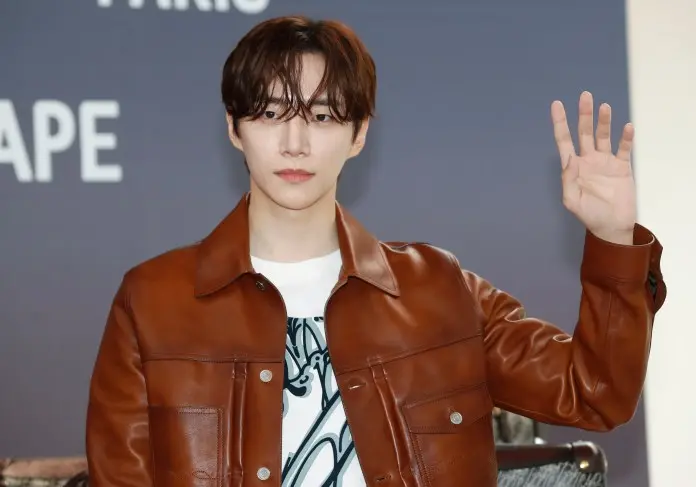Lee Junho with medium-length dark hair wearing a brown leather jacket and a white T-shirt, waving their hand while seated in front of a neutral background.