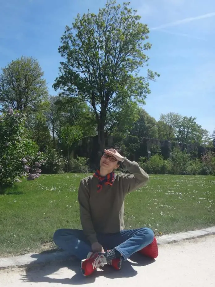 Song Hye-kyo cross-legged on the ground in a park, wearing casual clothing and a scarf, shading their eyes from the sun. Green trees and grass are visible in the background under a clear blue sky.