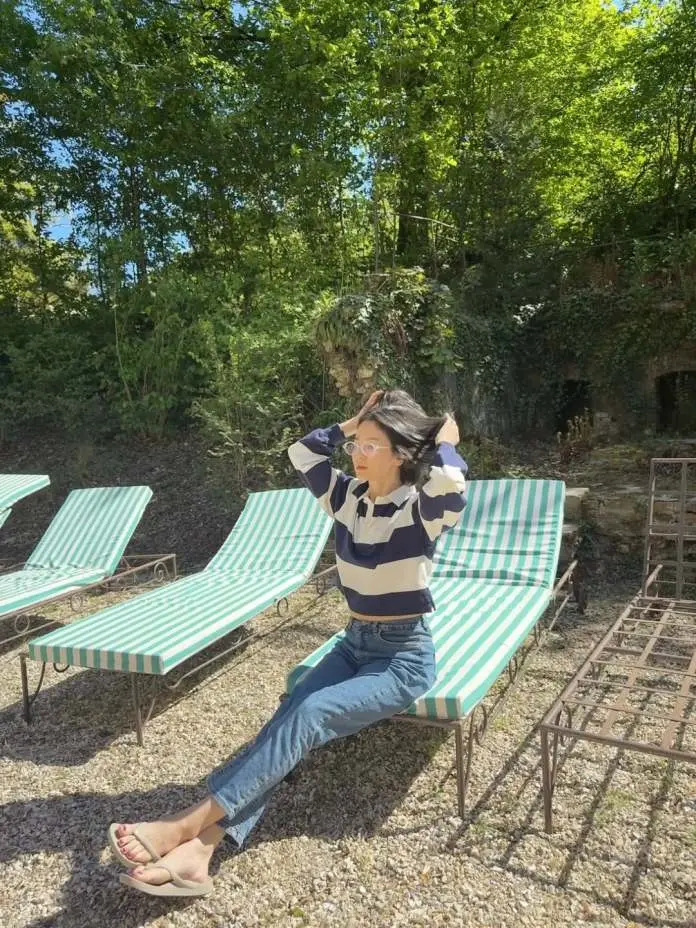 Song Hye-kyo sitting on a lounge chair in a sunny outdoor setting, wearing a striped long-sleeve top and blue jeans, with greenery in the background.