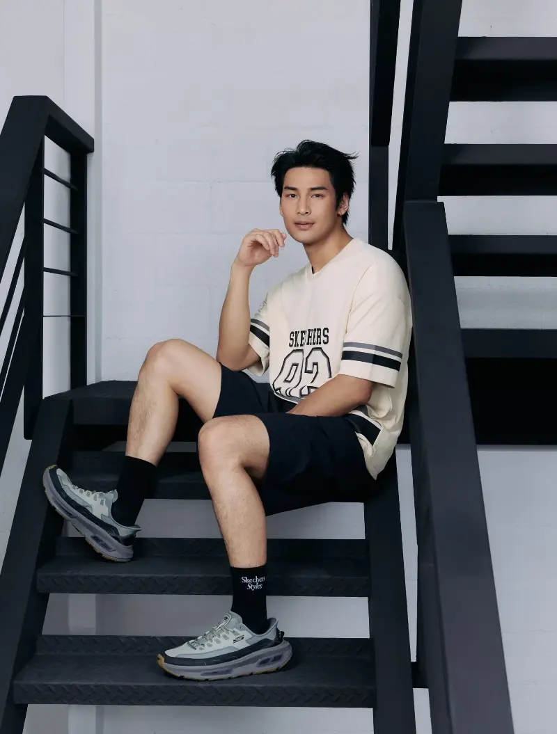 A man sits on black steel stairs in a cream Skechers tee, black shorts, and grey D'Lites 330 sneakers