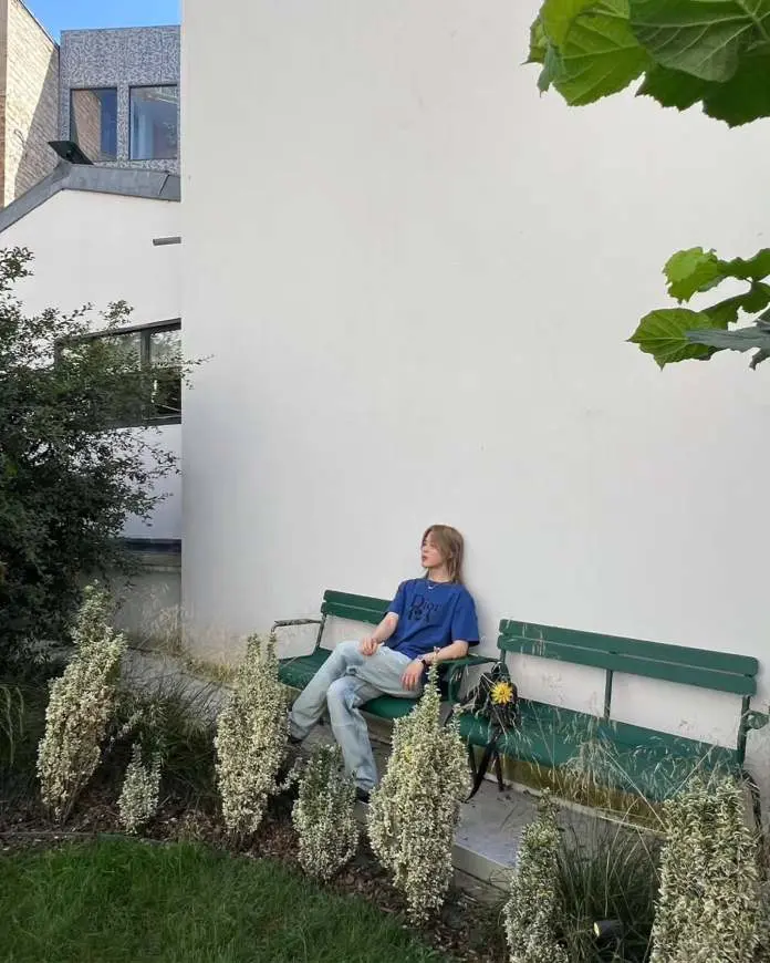 Jimin sitting on a green bench next to decorative plants in a minimalist outdoor setting, with a white wall and modern architecture in the background.