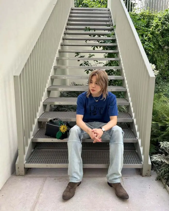 Jimin sitting on stairs surrounded by greenery, wearing a blue Dior t-shirt and light jeans, with a black bag and a sunflower.
