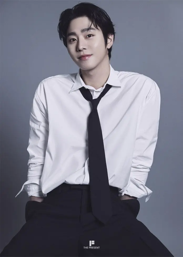 A young man with dark hair, wearing a white shirt and black tie, smiles at the camera against a grey background.