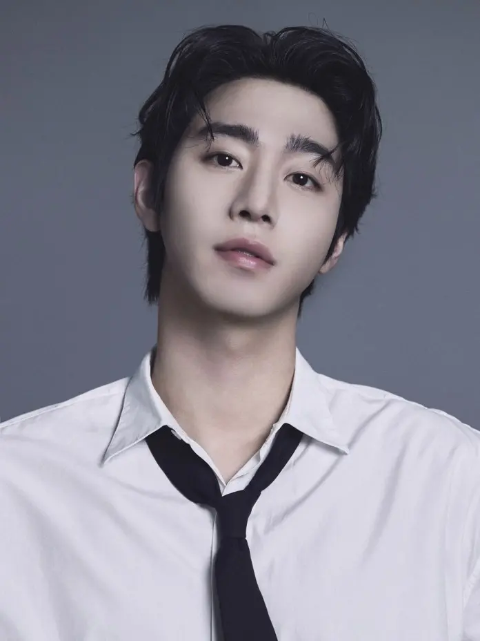 A young man with dark hair and a slight smile, wearing a white collared shirt and a black tie, against a gray background.