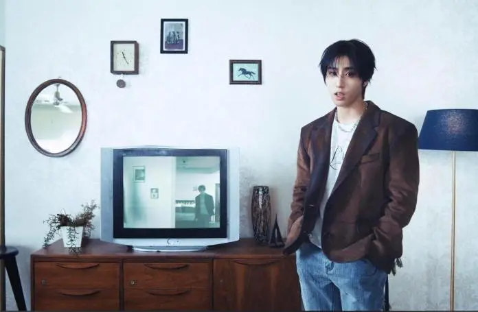 Han standing in a retro-styled living room, wearing a brown blazer and casual attire, with a vintage television displaying a reflection of another person. The room features patterned wallpaper, a mirror, and various framed pictures on the wall.