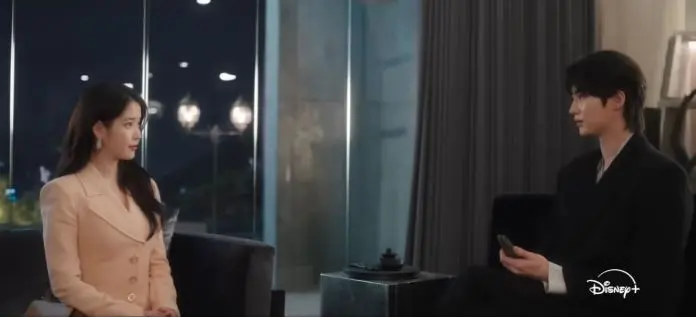 IU in a beige suit and Byeon Woo-seok in a black outfit sit across from each other in a well-lit room, engaged in conversation.