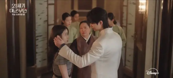 A dramatic scene featuring Byeon Woo-seok in a white suit holding the face of IU in a checkered dress, with a serious expression. In the background, two women in traditional attire observe the interaction.
